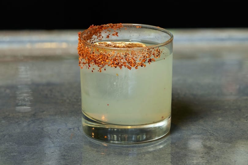 A light-colored cocktail in a glass with a chili-salt rim and a lime slice.