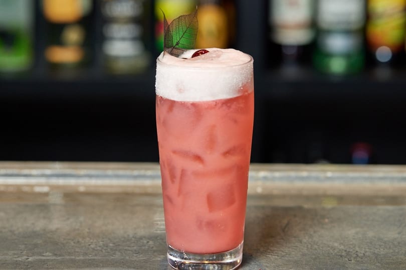 Pink cocktail with ice, foamy top, sugar rim, and a leaf garnish on a bar counter.