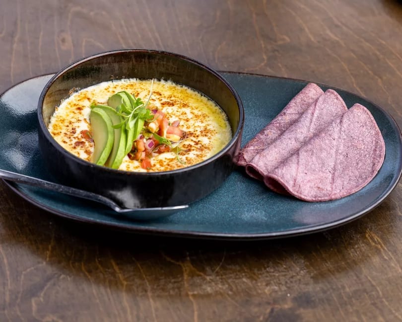 Melted cheese dip with avocado, pico de gallo, and purple tortillas on a dark plate.