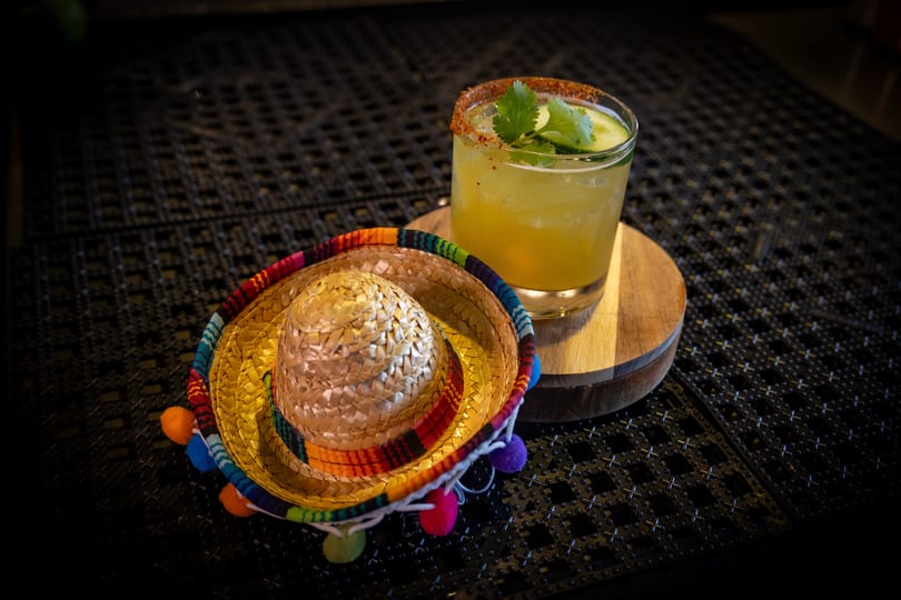 A cucumber margarita with a chili-salt rim next to a miniature sombrero on a dark textured surface.