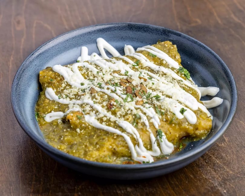 Green enchiladas topped with crema, cotija cheese, and pumpkin seeds in a dark blue bowl.