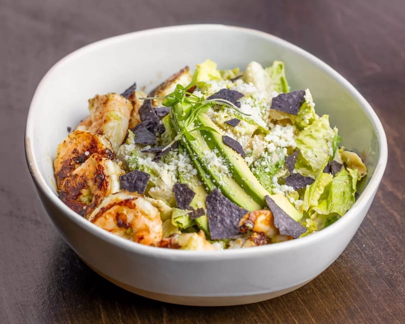 Grilled shrimp salad with avocado, blue tortilla chips, and crumbled cheese in a white bowl.