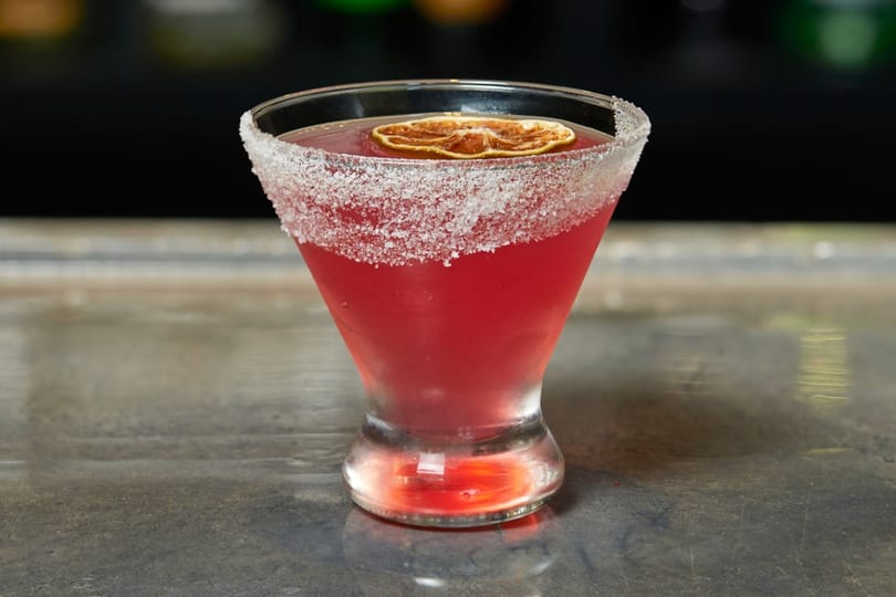 A vibrant red cocktail in a sugar-rimmed martini glass, garnished with a dried orange slice.