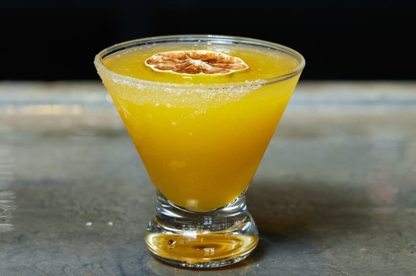 A vibrant yellow cocktail in a martini glass, garnished with a dried citrus slice and sugar rim.