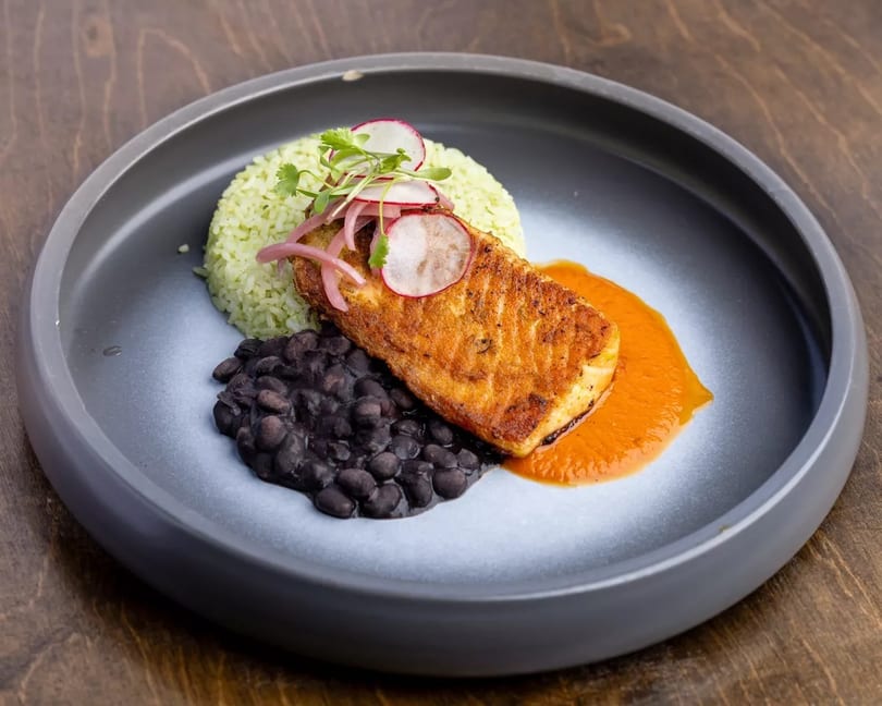 Pan-seared fish with green rice, black beans, and red sauce, garnished with radishes and herbs.